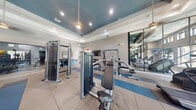 This generously sized fitness center is thoughtfully designed to accommodate a wide range of workout styles across a single open floor. The space is divided into distinct functional zones while maintaining an open, airy feel enhanced by elevated tray ceilings painted in a bold teal-blue accent, recessed lighting, and decorative pendant fixtures throughout.

The cardio zone features multiple treadmills, ellipticals, and stationary bikes positioned along large windows that offer views of an outdoor pool area, flooding the space with natural light. A wall-mounted television provides entertainment during workouts. The strength training area includes cable machines, a functional trainer, and a tiered dumbbell rack, all arranged on durable tile and rubberized flooring with blue accent strips.

A glass-enclosed studio room, framed by sleek black metal partitions, houses spin bikes, a heavy bag, medicine balls, kettlebells, plyo boxes, battle ropes, and a large-format display screen—ideal for group or high-intensity training. Full-length mirrors throughout amplify the sense of space and assist with form. The overall ambiance is modern and energetic, with a clean color palette of white, gray, and blue that contributes to a polished, professional fitness environment.