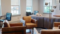 Located at 2674 Wildberry Ct in Edison, NJ, this single-floor living room offers a well-appointed, contemporary gathering space designed with both comfort and style in mind. The room features rich hardwood flooring throughout, complemented by a striking tray ceiling adorned with a multi-bulb statement chandelier that serves as a focal point overhead. A textured stone accent wall anchors the space, housing a sleek linear electric fireplace below a wall-mounted flat-screen TV, flanked by framed geometric artwork that adds a bold, modern touch. Warm wood shelving runs along the fireplace surround, adding a natural contrast to the light stone finish. Seating is thoughtfully arranged to encourage conversation and relaxation, with a large channeled gray sectional sofa accented by colorful patterned throw pillows, and a set of cognac leather arm chairs with industrial-style metal frames. A cluster of mixed cylindrical ottomans provides flexible additional seating. Multiple windows dressed in layered roman shades and roller blinds flood the room with natural light while maintaining privacy. A dedicated bistro-style table with chairs near the windows creates an intimate nook for casual use. The overall ambiance is modern and inviting, blending warm tones with contemporary finishes throughout.