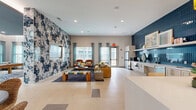 This vibrant communal living space is thoughtfully designed to blend comfort, function, and contemporary style across a single, open floor plan. The room is immediately defined by two striking accent walls — a bold, abstract-patterned wallcovering in navy, blush, and cream anchors the main lounge area and frames a wall-mounted flat-screen television, while a full-height, deep-blue vertical tile backsplash commands attention in the kitchenette zone. Large-format neutral floor tiles run throughout, contributing to a clean and cohesive aesthetic. Abundant natural light pours in through multiple oversized windows dressed with sheer teal curtains, offering views of the surrounding building exterior. The lounge area is furnished with a generous sectional sofa, a pair of rust-orange accent chairs, and a woven coffee table atop a navy area rug, creating an inviting gathering space. The integrated kitchenette features light wood cabinetry, stainless steel appliances including a refrigerator and dishwasher, open display shelving, and a white island with bar seating illuminated by pendant lighting with gold accents. Recessed ceiling lighting ensures the entire space is well-lit, while the open layout allows for flexible use as both a social lounge and functional amenity area.