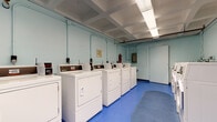 This shared laundry facility is designed for practicality and efficiency, offering a well-organized layout across a single level. The main area houses two rows of white top-load and front-load washing machines along with matching dryers, providing ample capacity for multiple users simultaneously. The machines are equipped with coin-operated or card-reader controls, making transactions straightforward. A utility sink station is positioned along one wall, offering additional washing or rinsing options.

The space features bright blue vinyl flooring throughout, which is both durable and easy to maintain. A gray oval floor mat is centrally placed, adding a practical touch. The walls are painted in a light blue tone, contributing to a clean and open atmosphere. Fluorescent lighting panels are recessed into a coffered ceiling, ensuring the entire room is well-lit.

An adjoining area provides extra functionality, featuring a dedicated folding table for sorting and folding laundry, along with additional dryer units and a wall-mounted fan for ventilation. A bulletin board displays facility rules and notices. Safety is addressed with a clearly visible fire extinguisher. The overall environment is utilitarian, clean, and efficiently arranged to serve the needs of multiple users.