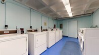 This shared laundry facility is designed for practicality and efficiency, offering a well-organized layout across a single level. The main area houses two rows of white top-load and front-load washing machines along with matching dryers, providing ample capacity for multiple users simultaneously. The machines are equipped with coin-operated or card-reader controls, making transactions straightforward. A utility sink station is positioned along one wall, offering additional washing or rinsing options.

The space features bright blue vinyl flooring throughout, which is both durable and easy to maintain. A gray oval floor mat is centrally placed, adding a practical touch. The walls are painted in a light blue tone, contributing to a clean and open atmosphere. Fluorescent lighting panels are recessed into a coffered ceiling, ensuring the entire room is well-lit.

An adjoining area provides extra functionality, featuring a dedicated folding table for sorting and folding laundry, along with additional dryer units and a wall-mounted fan for ventilation. A bulletin board displays facility rules and notices. Safety is addressed with a clearly visible fire extinguisher. The overall environment is utilitarian, clean, and efficiently arranged to serve the needs of multiple users.