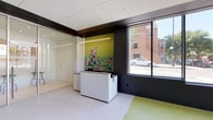 This single-floor commercial space is thoughtfully designed with a clean, contemporary aesthetic tailored for pet services. The reception area welcomes visitors with a sleek white front desk, a vibrant large-scale mural of a colorful dog, and a prominent picture window offering views of the surrounding streetscape. A bold contrast of matte black trim against white ceilings and walls defines the architectural language throughout.

The main open area features a soft green rubberized floor that transitions organically into a white textured surface, creating distinct activity zones without hard boundaries. A full wall clad in artificial turf adds a playful, nature-inspired element and provides functional texture. Large floor-to-near-ceiling windows span multiple walls, delivering abundant natural light and panoramic views of the urban neighborhood outside.

A glass-partitioned room with frosted panels sits adjacent to the main area, offering a semi-private space furnished with bar-height seating, ideal for consultations or staff use. Recessed ceiling lighting supplements the generous natural light throughout. The overall design balances a professional, modern commercial environment with a fun, animal-friendly atmosphere.