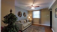 This two-story home offers a thoughtfully designed layout that balances comfort and functionality across both floors. The living room serves as a welcoming focal point, featuring warm neutral-toned walls that create a cozy atmosphere. A ceiling fan with a built-in light fixture anchors the room, providing both air circulation and ambient lighting. Natural light filters in through a window dressed with elegant grommet-top curtains, brightening the space with a soft, airy feel. Hardwood-style flooring extends throughout, complemented by a plush area rug that adds texture and warmth underfoot. Decorative sunburst wall art lends a stylish, contemporary accent to the living area, while lush indoor greenery adds a natural, refreshing touch. The overall design aesthetic is neutral and inviting, with a palette of warm taupes and soft grays that lend themselves to a variety of décor styles. With two floors of living space, this home provides ample room for both everyday living and entertaining, making it a versatile and practical residence suited to a range of lifestyles.