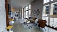 This single-floor space showcases a contemporary open-concept design characterized by dramatic double-height ceilings and an expansive, airy atmosphere. Polished concrete floors run throughout, lending an industrial yet refined aesthetic that complements the warm, neutral-toned walls. Floor-to-ceiling windows along the perimeter allow abundant natural light to pour into the interior while offering views to the outdoors.

The main lounge area is anchored by a patterned chevron sofa paired with a mid-century modern coffee table, all set atop a textured area rug. A cognac leather accent chair adds a warm contrast to the cool palette. Diamond-shaped framed art pieces adorn one wall, contributing a geometric focal point to the seating arrangement.

Structural columns are incorporated throughout the space, reinforcing the architectural character without interrupting the open flow. Toward the rear, a separate area with bar-height seating and stools is visible, suggesting a secondary gathering or social zone. Recessed lighting supplements the natural light, ensuring the space remains well-lit and inviting. The overall ambiance is modern, sophisticated, and welcoming.