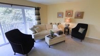 This single-floor residence offers a comfortable and thoughtfully designed living environment with a warm, inviting ambiance throughout. The living room serves as the heart of the space, showcasing soft yellow walls that lend a bright, cheerful atmosphere. Plush wall-to-wall carpeting adds a sense of coziness underfoot, while a large window dressed with bold plaid drapes frames a view of lush greenery beyond the private balcony, allowing natural light to fill the room. The living area is well-appointed with a cream-toned sofa, a cushioned ottoman with nailhead trim that doubles as a coffee table, and a pair of dark accent chairs that provide an elegant contrast. A classic table lamp with a warm glow and curated wall art add refined decorative touches. The balcony access further extends the living space outdoors, offering a peaceful retreat surrounded by nature. Overall, the space presents a harmonious blend of comfort and understated elegance, making it ideal for both relaxed everyday living and casual entertaining.