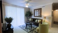 This single-floor residence centers around a thoughtfully designed living area that balances comfort and functionality. The living room is highlighted by elegant crown molding along the ceiling perimeter, adding a refined architectural detail to the space. A ceiling fan with integrated lighting is centrally positioned, providing both air circulation and ambient illumination.

Natural light is a prominent feature, courtesy of a large sliding glass door fitted with full-length vertical blinds, allowing for flexible light control and privacy. The carpeted flooring adds warmth underfoot and is complemented by a patterned area rug that anchors the seating arrangement.

The living area is generously proportioned, comfortably accommodating a plush sofa adorned with decorative pillows, a cushioned accent chair, and a round glass-top coffee table. A tall indoor plant introduces a natural, organic element to the interior, while a framed wall decoration and stylish table lamp contribute to the overall polished aesthetic. The neutral wall tones and coordinated furnishings create a cohesive, inviting atmosphere throughout the space.