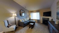 This single-floor residence offers a comfortable and well-appointed living space designed with both style and functionality in mind. The living room serves as the heart of the home, featuring vaulted ceilings that lend an airy sense of openness to the space. Soft, neutral carpeting runs throughout, contributing to a warm and inviting atmosphere.

The room is anchored by a sectional sofa in a cool slate blue, accented with floral throw pillows that add a touch of color and character. A decorative ornate mirror with an antique-style frame adorns the blue-gray accent wall, complementing the warm yellow tones of the adjacent walls. Natural light filters in through two windows dressed in patterned sheer curtains, brightening the space throughout the day.

A fireplace with a clean, tiled surround serves as a focal point on one side of the room, providing both visual interest and a source of warmth. A wall-mounted flat-screen television is positioned nearby for convenient viewing. Nesting side tables and a classic floor lamp with a turned wooden base complete the thoughtfully curated living area. Overall, the space presents a balanced blend of comfort and understated elegance, making it a functional and welcoming home environment.