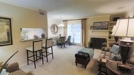 This single-floor space showcases an open-concept layout that seamlessly connects the living, dining, and kitchen areas. The living room is anchored by a classic fireplace with a decorative wrought-iron screen and a traditional white mantle, adding both warmth and character to the room. Soft, neutral-toned carpeting runs throughout, contributing to a comfortable and cohesive aesthetic.

A breakfast bar with bar-height seating provides a casual dining option and serves as a natural divider between the kitchen and the main living area. Adjacent to this, a dedicated dining nook features elegant upholstered chairs and is enhanced by a pendant chandelier overhead and a decorative sunburst mirror on the wall.

Natural light fills the space through a large window dressed with white plantation shutters and flowing gold drape panels, offering a bright and airy atmosphere. Crown molding details lend a refined architectural touch throughout. The overall ambiance is warm and inviting, with a neutral color palette of beige and cream tones that create a timeless, well-appointed interior.