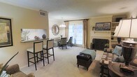 This single-floor space showcases an open-concept layout that seamlessly connects the living, dining, and kitchen areas. The living room is anchored by a classic fireplace with a decorative wrought-iron screen and a traditional white mantle, adding both warmth and character to the room. Soft, neutral-toned carpeting runs throughout, contributing to a comfortable and cohesive aesthetic.

A breakfast bar with bar-height seating provides a casual dining option and serves as a natural divider between the kitchen and the main living area. Adjacent to this, a dedicated dining nook features elegant upholstered chairs and is enhanced by a pendant chandelier overhead and a decorative sunburst mirror on the wall.

Natural light fills the space through a large window dressed with white plantation shutters and flowing gold drape panels, offering a bright and airy atmosphere. Crown molding details lend a refined architectural touch throughout. The overall ambiance is warm and inviting, with a neutral color palette of beige and cream tones that create a timeless, well-appointed interior.