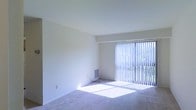 This single-floor space offers a clean, functional layout designed to maximize comfort and usability. The main living area is generously proportioned, featuring plush wall-to-wall carpeting that adds warmth underfoot. Crisp white walls and a smooth, high ceiling contribute to an open, airy atmosphere throughout the room.

A prominent sliding glass door serves as the focal point of the living area, fitted with full-length vertical blinds that allow for precise control of natural light. When the blinds are open, sunlight streams across the carpeted floor, brightening the entire space. The door also provides a view of the lush greenery outside, offering a pleasant outdoor connection.

A built-in wall vent indicates a dedicated HVAC system for year-round climate control. The room's neutral color palette and uncluttered design make it highly adaptable to a variety of furniture arrangements and interior styles. The overall ambiance is calm and welcoming, presenting a versatile canvas ready for personalization.