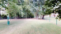 This dog park offers a welcoming and well-maintained outdoor environment designed for canine recreation and socialization. The grounds are anchored by a generous open lawn area, providing ample room for dogs to run, play, and explore freely. A perimeter fence — visible along the boundary — ensures a secure, contained space for off-leash activity.

Mature, full-canopy trees are distributed throughout the park, offering generous natural shade across much of the grounds. The tree coverage creates cooler, comfortable zones during warmer months and adds a pleasant, park-like atmosphere to the setting. Fallen leaves scattered across the grass suggest a lush, established landscape that changes naturally with the seasons.

A picnic table is situated beneath the tree canopy, providing a resting spot for owners while keeping watch over their pets. The overall layout balances open sunny areas with shaded retreats, catering to a variety of needs for both dogs and visitors.

The park has a natural, unhurried ambiance with a grassy surface underfoot and a relaxed, community-oriented character. Its straightforward, functional design prioritizes open space and safety, making it a practical and enjoyable destination for dog owners looking for a dedicated, secure outdoor area for their pets.