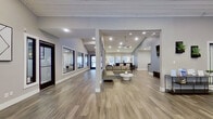This well-appointed single-story commercial space presents a thoughtfully designed open-concept layout that balances professional functionality with contemporary style. The entrance greets visitors through elegant dark-wood French doors, opening into a spacious foyer with vaulted, white-painted shiplap ceilings and recessed lighting that illuminate the warm-toned luxury vinyl plank flooring throughout.

The reception area features a dedicated workstation equipped with computer terminals and accompanied by modern cantilever chairs, creating an organized and welcoming point of contact. A sleek display console along the wall provides space for informational materials and decorative greenery, adding a polished finishing touch.

Adjacent to the reception is an open lounge area furnished with a generous neutral sectional sofa, a round white coffee table, and views into interior windowed rooms—likely private offices or conference spaces—that maintain visual connectivity while ensuring privacy. A separate, more intimate lounge area offers an orange accent sofa, patterned rug, and large shuttered windows that invite abundant natural light and outdoor views.

Structural columns define distinct zones throughout the space without interrupting the open flow. Cool gray walls, crisp white trim, and consistent flooring unify the interior, creating a clean, modern ambiance suited to a professional environment.