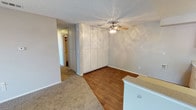 This single-floor residence presents a clean, functional design with a welcoming and straightforward layout. The main living area showcases a seamless transition between warm wood-style hard flooring and plush carpeting, creating distinct zones within the open space. A ceiling fan with a light fixture is centrally positioned, providing both air circulation and ambient lighting throughout the room.

A standout feature of the space is the floor-to-ceiling built-in cabinetry unit, which offers generous storage capacity and a streamlined, organized aesthetic. The textured popcorn ceiling adds a classic residential character, while the neutral gray walls provide a versatile backdrop suitable for a variety of décor styles.

A partial countertop or bar-height surface is visible toward the foreground, suggesting a convenient transitional space between the living and kitchen areas. A hallway leading further into the unit hints at additional rooms beyond, visible in the background with warm lighting. The space is practical and well-appointed, offering ample storage, durable flooring choices, and a neutral palette that makes it easy to personalize.
