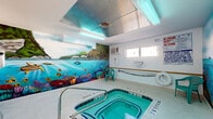 This indoor aquatic space is defined by its striking, full-wall tropical mural that commands immediate attention. The artwork depicts a vivid underwater reef scene complete with colorful fish, sea turtles, coral formations, and a lush island paradise above the waterline — creating an immersive, resort-like atmosphere within an enclosed environment. The centerpiece of the room is a tiled pool with clearly marked depth indicators along its edge, accompanied by stainless steel entry rails for safe and easy access. A dedicated hot tub sits adjacent, offering a relaxing complement to the main pool. The surrounding deck is finished in light-colored tile, providing a clean and functional surround. Seating is available poolside in the form of teal plastic chairs positioned along the walls. The room is illuminated by overhead fluorescent lighting, with supplemental natural light entering through high, horizontally oriented windows. A wall-mounted air conditioning unit ensures climate control year-round. Safety is a clear priority, with visible life rings, caution signage, an emergency shut-off switch, and pool rules posted throughout. The ceiling features a partial metallic panel detail above the pool area, adding an architectural accent. An exit door is accessible directly from the pool room, ensuring convenient egress.