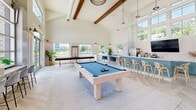 Located at 210 Brunswick Pl in Myrtle Beach, SC, this single-floor community clubhouse is a beautifully designed gathering space that blends coastal charm with modern amenities. The interior is defined by dramatic vaulted ceilings with exposed wooden beams that soar overhead, flooding the space with an open, expansive feel. Large clerestory windows and oversized glass doors draw in abundant natural light and frame views of the surrounding outdoor pool area, seamlessly connecting the indoors with the outdoors.

The living area features a sophisticated coastal aesthetic, with shiplap-style white panel walls, light herringbone-pattern hardwood flooring, and a wall-mounted flat-screen television above a warm wood credenza. Woven rattan accent chairs, a plush sofa, and layered area rugs create an inviting lounge setting complemented by lush indoor greenery.

The space transitions into a dynamic recreation zone anchored by a striking white billiard table with a blue felt surface, pendant lighting above, and a full-length shuffleboard table positioned along the perimeter. A teal-accented wet bar with bar stool seating provides a social hub, while double glass doors open directly to the outdoor pool deck. The overall ambiance is bright, welcoming, and thoughtfully designed for community enjoyment.