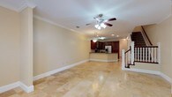 Located at The Gardens, this two-story residence offers a thoughtfully designed layout that balances comfort and functionality. The main level showcases a spacious open-plan living area finished with large-format polished travertine tile flooring that reflects light beautifully across the room. Crown molding runs throughout, lending an elegant, refined touch to the interior, while a ceiling fan with integrated lighting ensures year-round comfort.

The living area flows seamlessly into a well-equipped kitchen, which features rich dark wood cabinetry, dark stone countertops, and a central island that serves as both a workspace and a casual gathering spot. Stainless steel appliances, including a French door refrigerator, add a modern, polished finish to the culinary space. Recessed lighting overhead keeps the kitchen bright and functional.

A classic staircase with white balusters and a dark wood handrail anchors the main level and leads to the second floor, adding a traditional architectural element to the home. The overall ambiance of the space is bright, clean, and inviting, with a neutral color palette that provides a versatile backdrop. This home at The Gardens combines timeless design details with practical, modern amenities across both levels.