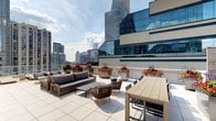 This rooftop terrace offers an expansive open-air retreat set against a striking urban backdrop. The space is finished with large-format light-toned pavers that lend a clean, contemporary aesthetic underfoot. A comfortable lounge area anchors one side of the terrace, featuring a generously sized sofa with deep charcoal cushions, paired with woven accent chairs and a low-profile coffee table — all constructed from natural wood tones that complement the outdoor setting. On the opposite side, a sizable rectangular dining table with matching chairs provides an ideal setting for al fresco gatherings. Vibrant planters filled with colorful seasonal flowers — including rich reds, oranges, and purples — are positioned throughout the terrace, adding lively bursts of color against the neutral palette. The surrounding cityscape forms a dramatic backdrop, with mid- and high-rise buildings visible in multiple directions, including a prominent glass curtain-wall tower that reflects the sky. The terrace benefits from abundant natural daylight and open sky views, creating a bright and energizing atmosphere well-suited for both casual relaxation and entertaining.