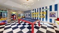 Located at 2150 Vining Dr in Woodbury, MN, this energetic game room is a single-floor entertainment space designed to impress. The room makes an immediate visual statement with its bold black-and-white checkered flooring, vivid blue-and-white striped accent wall, and eye-catching gold "GAME ON PLAY ON" lettering. A striking modern chandelier composed of interlocking rings hangs overhead, casting warm light throughout the space and complementing the recessed lighting above.

At the center of the room sits a red-framed billiard table with a navy felt surface, serving as the focal point of the space. Along one side, a dedicated arcade zone features classic cabinet games including a racing simulator, a basketball shooting game, and a Pac-Man cabinet, all set against a colorful comic book-themed wallpaper that adds a playful, retro energy.

Natural light filters in through multiple white-framed windows, keeping the space bright and inviting. A built-in bench with a yellow cushion and red accent pillows provides casual seating near an arched window nook. A glass partition separates the game area from a bright, adjacent office workspace with hardwood flooring and a clean white desk, offering a seamless blend of recreation and function.