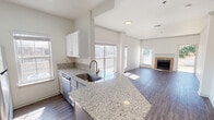 This single-floor residence offers a well-designed, open-concept layout that seamlessly connects the kitchen and living areas. The kitchen is a standout feature, showcasing granite countertops with a large peninsula that doubles as a breakfast bar, an undermount sink with a brushed gold faucet, white shaker-style cabinetry, a classic subway tile backsplash, and stainless steel appliances including a range, microwave, and refrigerator. A skylight above the kitchen adds a bright, airy feel to the space. The living area is generously proportioned, filled with natural light from multiple large windows, and anchored by a tile-surround fireplace that serves as a focal point. Dark hardwood-style flooring runs throughout the main living spaces, complementing the neutral white walls and crown molding details. The bedroom features plush carpet flooring, a ceiling fan, and a window offering outdoor views. The bathroom is appointed with granite-topped grey vanity cabinetry, a tiled soaking tub and shower combination with a distinctive red accent tile border, and wood-style flooring. Throughout the residence, clean lines, neutral tones, and consistent finishes create a cohesive and inviting atmosphere.