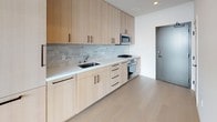 This thoughtfully designed single-floor residence combines clean modern aesthetics with functional living. Light wide-plank hardwood flooring runs throughout the main living areas, creating a warm, cohesive foundation that complements the crisp white walls and recessed lighting overhead.

The kitchen is outfitted with flat-front light wood cabinetry extending to the ceiling for generous storage, paired with a stone mosaic tile backsplash and white countertops. Stainless steel appliances — including a gas cooktop, oven, and built-in microwave — complete the well-equipped cooking space. Dark hardware accents provide a refined contrast throughout.

The open living area flows naturally from the kitchen and hallway, bathed in abundant natural light. A dramatic floor-to-ceiling window wall with a glass-panel balcony door anchors the space, framing expansive panoramic city views that stretch across the skyline — a standout feature of this residence.

The bathroom is finished with large-format floor tile and a frameless glass-enclosed walk-in shower clad in textured stone mosaic tile, lending a spa-like quality to the space. A vanity and modern fixtures complete the room.

The entry corridor connects all areas efficiently, with a neutral palette and recessed lighting maintaining a consistent, contemporary feel throughout the entire unit.