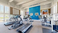 Located at 2515 Northeast Expressway in Atlanta, GA, this fitness center offers a generously sized, single-floor workout facility designed to support a full range of exercise routines. The space is defined by its impressively high ceilings with exposed beam detailing, which lend an open and airy atmosphere throughout.

The main workout area is thoughtfully arranged with a variety of cardio equipment, including treadmills, elliptical trainers, and a stationary bike, as well as strength training machines and open floor space equipped with exercise mats. A bold blue accent wall provides a vibrant focal point, complemented by framed cityscape artwork that adds a modern, urban aesthetic. Wall-mounted televisions are positioned for convenient viewing during workouts.

Full-length mirrors line the walls, enhancing the sense of space and allowing users to monitor their form. Large, multi-pane windows flood the interior with natural light and offer views of the surrounding outdoor greenery. An elegant staircase with wrought iron railings is visible from the main floor, adding an architectural touch.

The entry area includes a vending machine and seating, providing convenience for visitors. Carpeted flooring with protective rubber mats beneath equipment ensures comfort and safety. The overall ambiance is clean, bright, and motivating — a well-maintained facility suited for a comprehensive fitness experience.
