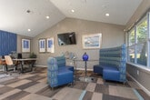 This single-floor community clubhouse and leasing office offers a well-designed, open-concept layout that balances functionality with a comfortable, inviting atmosphere. The space is anchored by a bold, multi-toned geometric carpet that defines distinct zones throughout the interior, adding visual interest and a modern flair underfoot.

The main lounge area features vaulted ceilings with exposed wood beam accents, recessed lighting, and elegant globe-style chandeliers that cast a warm glow over the seating arrangements. Furnishings include patterned yellow armchairs, blue accent chairs, and side tables, creating an approachable and stylish gathering space. A wall-mounted flat-screen television and framed artwork lend the area a polished, residential feel.

Large windows along multiple walls flood the interior with abundant natural light and provide views of the lush greenery and outdoor pool area. Glass slider doors lead directly to the pool, seamlessly connecting the indoor and outdoor environments.

The leasing desk area is positioned near the main entrance, offering a professional yet welcoming reception point with seating for visitors. Adjacent private offices enclosed with glass-panel partitions provide quieter, dedicated workspaces. High-top workstations with computers are also available for resident use. Potted plants are thoughtfully placed throughout, adding a touch of life and warmth to the overall space.