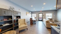 Located at 2106 Runestone Ave in Alexandria, Minnesota, this single-floor communal space offers a practical and welcoming open-plan layout that seamlessly integrates a living room and dining area. The interior is finished with durable wood-look plank flooring throughout, smooth gray-toned walls, and recessed lighting that provides consistent, even illumination across the space.

The dining area is anchored by a striking solid wood table with richly grained matching chairs, accompanied by a wall-mounted ductless HVAC unit for year-round climate control. Natural light filters in through multiple windows, and a door provides direct access to an outdoor area. A colorful floral canvas print adds a warm accent to the neutral wall.

The living area features a comfortable neutral-toned sofa and accent chairs arranged around rustic wood-and-metal coffee tables, with a wall-mounted flat-screen television as a focal point. Baseboard heating runs along the perimeter, supplementing the ductless system.

A compact kitchenette occupies one corner, equipped with gray cabinetry, a sink, countertop appliances including a coffee maker and microwave, a dishwasher, and a full-size refrigerator — providing functional amenities for group use. A storage unit with cabinetry offers additional organizational space. The overall design balances modern, clean aesthetics with warm, natural material accents, creating a comfortable and functional shared environment.