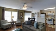 This single-floor residence offers a thoughtfully designed open-concept layout that seamlessly connects the living, dining, and kitchen areas into one cohesive and functional living space. The living room serves as the heart of the home, featuring warm hardwood-style flooring, earthy olive-toned accent walls, and a ceiling fan with integrated lighting that adds both comfort and style. Large windows dressed with vertical blinds and decorative curtains allow generous amounts of natural light to fill the space, creating a bright and welcoming atmosphere. The seating area is well-proportioned, comfortably accommodating a sofa and accent chair, while a glass-top coffee table adds a contemporary touch. Adjacent to the living area, the dining space features a compact table and chairs, ideal for everyday meals and casual gatherings. Built-in shelving units frame the dining area, providing both display and storage opportunities. The kitchen is partially visible beyond the dining space, featuring white cabinetry and appliances, including a full-sized refrigerator, connected via a pass-through counter that enhances the open feel of the floor plan. The textured ceiling throughout adds a classic residential character to the overall interior design.
