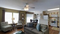 This single-floor residence offers a thoughtfully designed open-concept layout that seamlessly connects the living, dining, and kitchen areas into one cohesive and functional living space. The living room serves as the heart of the home, featuring warm hardwood-style flooring, earthy olive-toned accent walls, and a ceiling fan with integrated lighting that adds both comfort and style. Large windows dressed with vertical blinds and decorative curtains allow generous amounts of natural light to fill the space, creating a bright and welcoming atmosphere. The seating area is well-proportioned, comfortably accommodating a sofa and accent chair, while a glass-top coffee table adds a contemporary touch. Adjacent to the living area, the dining space features a compact table and chairs, ideal for everyday meals and casual gatherings. Built-in shelving units frame the dining area, providing both display and storage opportunities. The kitchen is partially visible beyond the dining space, featuring white cabinetry and appliances, including a full-sized refrigerator, connected via a pass-through counter that enhances the open feel of the floor plan. The textured ceiling throughout adds a classic residential character to the overall interior design.