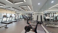 This modern fitness center in Addison, Texas, offers a thoughtfully designed workout space that maximizes both functionality and comfort. The open floor plan accommodates a comprehensive range of equipment, creating distinct zones for cardio, strength training, and functional fitness without feeling cramped.

The cardio section lines the perimeter and features multiple True treadmills and elliptical machines positioned in front of large windows fitted with roller shades, allowing natural light to filter in while offering views of the landscaped outdoor surroundings. This placement creates an energizing environment for endurance workouts.

A robust strength training area includes a multi-functional cable machine, a full rack of dumbbells with accompanying benches, and a selection of weighted bars and plates. Peloton spin bikes with interactive screens are centrally positioned, catering to high-intensity cycling sessions. Additional functional fitness equipment such as medicine balls, stability balls, and rowing machines round out the offerings.

Floor-to-ceiling mirrors line multiple walls, visually expanding the space and allowing users to monitor their form throughout their workouts. Overhead, a distinctive coffered ceiling features linear LED lighting panels and ceiling fans, providing consistent, bright illumination throughout the facility. Durable rubber flooring covers the entire space, ensuring safety and comfort. The entrance features glass double doors, contributing to the clean, contemporary aesthetic of this well-appointed fitness facility.