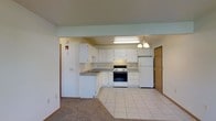 This single-floor space showcases a functional and well-organized layout, with an open-concept kitchen that flows naturally into the adjacent living area. The kitchen is outfitted with ample white cabinetry offering generous storage space, complemented by a full suite of white appliances, including a refrigerator, electric range with oven, and a dishwasher. A built-in microwave or range hood is positioned above the cooktop, adding to the kitchen's practicality. The countertops provide a workable surface area, and a sink is conveniently situated beneath the upper cabinetry. A chandelier-style light fixture and a recessed fluorescent light illuminate the kitchen, creating a bright and welcoming atmosphere. The kitchen floor features classic white square tiles, which transition into carpeting in the living area, defining each zone while maintaining a cohesive feel throughout the space. Neutral wall tones lend a clean, understated aesthetic throughout. A doorway visible at the rear of the space leads to additional rooms, suggesting a practical and well-connected floor plan. The overall ambiance is clean, bright, and move-in ready.