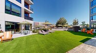 Located at 377 Crane St in Orange, NJ, this outdoor amenity space is a thoughtfully designed courtyard that serves as a central gathering hub for the surrounding multi-story building. The grounds feature expansive, well-maintained artificial turf lawns that create a lush, verdant setting beneath open skies, bathed in abundant natural light throughout the day.

The courtyard is anchored by multiple distinct seating areas, including vibrant orange Adirondack chairs arranged around a central gathering point, as well as sleek lounge chairs paired with green market umbrellas for shaded relaxation. Stone-accented retaining walls add a natural, textural element to the landscape, while oversized decorative planters — some filled with flowering plants and small trees — punctuate the space with greenery and visual interest.

Manicured garden beds line portions of the perimeter, featuring ornamental grasses, seasonal flowers, and shrubs that soften the modern architectural lines of the surrounding building. The building itself presents a contemporary facade with large windows, private balconies, and warm wood-toned accents that frame the courtyard on multiple sides, creating an intimate yet open-air feel. Paved walkways connect the various zones seamlessly, offering easy flow throughout the space. This courtyard delivers a resort-inspired outdoor experience within an urban setting.