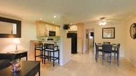 This well-appointed single-floor space offers an open, airy layout that connects the kitchen, dining, and living areas in a cohesive and functional flow. The kitchen is outfitted with light maple cabinetry, recessed lighting, black appliances including a full-size refrigerator and microwave, and a breakfast bar with seating for casual dining. The countertops provide generous prep space, and the cabinetry extends to the ceiling for ample storage. A partial dividing wall separates the kitchen from the adjacent dining area without closing off the open feel of the space. The dining area accommodates a full table and chairs, illuminated by a ceiling fan with integrated lighting that adds both comfort and practicality. Light hardwood flooring runs throughout, lending warmth and visual continuity to each area. Crown molding adds a refined architectural detail to the interiors, while neutral wall tones create a clean, versatile backdrop. Natural light enters through a window visible at the end of the hallway, brightening the space further. The layout is designed for ease of movement and everyday living, making it both welcoming and highly functional.