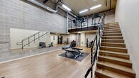 Located at 390 E Main St in Hillsboro, Oregon, this two-level exercise facility offers a well-appointed fitness environment designed for a comprehensive workout experience. The main floor is defined by impressively high ceilings and an open, airy layout, with light hardwood-style flooring throughout that lends a clean, modern aesthetic. Exposed concrete block walls and industrial-style ductwork overhead contribute a distinct urban character to the space. Large full-wall mirrors line one side of the main floor, visually expanding the room and supporting functional training. The main level accommodates free weights, a dumbbell rack, adjustable benches, and open floor space for stretching and mat work, all illuminated by bright overhead fluorescent lighting. A sleek staircase with a modern metal railing and cable detailing leads to the mezzanine level, which overlooks the main floor and houses additional cardio machines, including upright equipment and a treadmill. The lower mezzanine area beneath the staircase provides space for a cable machine, stability equipment, and supplemental fitness accessories. A glass entry door at the front of the facility allows natural light to filter in while offering views of the surrounding streetscape. This thoughtfully designed, multi-level fitness space provides a functional and motivating environment suited for a wide variety of exercise routines.