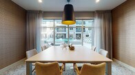 This single-floor business center is thoughtfully designed to support a variety of professional needs in a polished, contemporary setting. The space is anchored by a well-appointed main conference room featuring a large rectangular table surrounded by stylish upholstered chairs with gold-toned legs. A striking oversized black pendant light with a warm gold interior serves as a focal point, while expansive floor-to-ceiling windows dressed with sheer and blackout curtains flood the room with natural light and offer views of an outdoor pool courtyard. A textured herringbone-patterned accent wall adds warmth and visual depth to the conference area. Throughout the space, patterned carpeting in soft neutral tones provides a cohesive and professional aesthetic. Private workstation alcoves, painted in a calming blue-gray hue, feature built-in floating desks accented with warm wood edges and decorative gold wall art, offering focused individual work areas. A dedicated micro conference room provides an intimate setting for small meetings or calls, identified by clear signage and accessed through a distinctive perforated-panel door. The entry corridor leads visitors seamlessly through the space, with curated artwork and warm accent walls contributing to a refined and welcoming atmosphere throughout.