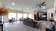 This single-floor space offers a generously proportioned open-plan layout that seamlessly blends a lounge area with a casual dining section. The living area is anchored by a large, plush sectional sofa in charcoal gray, complemented by a decorative patterned area rug and a red accent chair, creating a welcoming and stylish gathering space. A dark entertainment console with a wall-mounted television serves as a focal point on one side, flanked by decorative vases and sculptural accents that add an artistic touch to the room.

The dining area is positioned near a bank of large windows dressed with white blinds, allowing an abundance of natural light to fill the space. Multiple dining tables with mixed seating arrangements provide ample room for communal use and socializing.

Throughout the space, neutral beige walls and patterned carpet flooring create a cohesive and understated aesthetic. Multiple ceiling fans are installed across the room, enhancing airflow and comfort. Recessed lighting supplements the natural light, ensuring the space is well-illuminated at all times. The overall ambiance is comfortable, functional, and thoughtfully appointed, making it well-suited as a shared lounge or clubhouse common area.