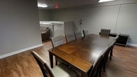 This single-floor space is designed to accommodate group gatherings and communal activities. The centerpiece of the room is a large, dark wood dining table with an inlaid geometric pattern, surrounded by upholstered tufted chairs in a neutral gray-green tone, offering seating for multiple guests. The hardwood-style flooring in warm brown tones adds a grounded, practical aesthetic throughout the space.

Adjacent to the dining area, a lounge section features a tan sofa, providing a comfortable secondary seating option. A partial half-wall with a white countertop ledge separates this area subtly, adding architectural definition without fully dividing the space.

The walls are finished in a muted gray tone, contributing to a neutral and functional atmosphere. Overhead fluorescent lighting ensures the space is well-lit, complemented by an illuminated exit sign and a visible fire extinguisher, indicating that the space meets standard safety requirements. The open layout allows for flexible use, whether for dining, meetings, or social gatherings, making this a versatile communal space.