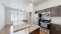 This well-appointed single-floor residence offers a thoughtfully designed layout that combines comfort and modern style. The heart of the home is an open-concept kitchen, anchored by a central island topped with granite countertops that provide generous prep and dining space. Rich, dark-toned cabinetry with sleek hardware lines the walls, complemented by a classic white subway tile backsplash. Stainless steel appliances — including a refrigerator, range, microwave, and dishwasher — complete the kitchen's contemporary feel. Pendant lights hang above the island, adding both task lighting and a stylish accent to the space. Large windows invite an abundance of natural light into the living area, which features warm hardwood-style flooring that extends throughout the main living spaces. The bedroom is finished with soft carpeting underfoot and offers convenient access to an in-unit stacked washer and dryer, adding a practical touch to everyday living. The bathroom mirrors the kitchen's refined aesthetic, showcasing matching granite countertops, dark cabinetry, and a tiled shower. Tall ceilings throughout the residence enhance the sense of openness, creating a bright and airy ambiance that makes this space both functional and inviting.