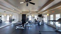 This fitness center offers a comprehensive workout environment designed to accommodate a range of exercise needs. The space is outfitted with a multi-station strength training machine, a weight bench, and an assortment of cardio equipment including an elliptical trainer and a stair climber, each equipped with individual display consoles. Dark rubber flooring provides a durable, non-slip surface suited for high-activity use throughout the room. A wall-mounted flat-screen television is positioned for easy viewing during workouts. Overhead, two ceiling fans help maintain air circulation, complemented by recessed lighting and a tray ceiling detail that adds an element of architectural refinement. Large windows along one wall allow ample natural light to fill the space while offering views to the outdoors, creating an open and energizing atmosphere. A wall-mounted equipment rack near the entrance provides additional storage and organization. The overall design strikes a balance between functionality and comfort, making this fitness center a practical and inviting destination for daily exercise routines.