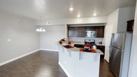 This well-appointed single-floor space showcases a thoughtfully designed open-concept layout that seamlessly connects the kitchen and dining areas. The kitchen is a standout feature, boasting rich dark-brown cabinetry that provides generous storage and a striking contrast against the light granite countertops. A peninsula with bar-height seating extends into the living space, creating a natural gathering spot and an ideal area for casual dining or entertaining. Stainless steel appliances, including a range and built-in microwave, add a modern touch to the kitchen. Recessed lighting throughout the kitchen keeps the space bright and functional, while a decorative chandelier over the dining area adds a touch of elegance. The flooring is a rich, dark hardwood-style material that runs continuously through the open-plan areas, lending warmth and cohesion to the space. Light gray walls and white trim contribute to a clean, contemporary aesthetic, keeping the atmosphere airy and inviting. Ample natural and recessed lighting ensures the space feels open and welcoming throughout the day. The overall design balances practicality with style, making it a comfortable and visually appealing living environment.