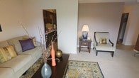 This single-floor residence offers a comfortable and cohesive living environment defined by warm, neutral tones and thoughtful design details throughout. The living area serves as the heart of the space, featuring plush carpeting layered with a decorative area rug in rich earth tones, creating an inviting and grounded atmosphere. Tasteful furnishings, including a cushioned sofa and an upholstered accent chair, are complemented by a dark wood coffee table and side table adorned with decorative vases and soft lamp lighting, lending the room a refined yet approachable character. A open passthrough connects the living area to the kitchen, which showcases dark wood cabinetry, a light-toned countertop, and a sink beneath overhead storage — offering both functionality and a striking contrast to the lighter tones of the main living space. The neutral wall palette throughout enhances the sense of openness and allows the décor and furnishings to take center stage. The overall ambiance of the space is warm, polished, and move-in ready, with a layout that promotes easy flow between the primary living and kitchen areas.