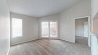 This single-floor space is thoughtfully laid out, offering a comfortable and functional living environment. The living area serves as the centerpiece of the home, featuring neutral-toned walls, plush wall-to-wall carpeting, and a textured ceiling that adds subtle visual interest. A set of French doors with gridded glass panels floods the room with natural light while providing access to the outdoors, creating an inviting and airy atmosphere. A secondary window along the adjacent wall further enhances the brightness of the space.

The entryway transitions seamlessly from hard-surface flooring into the carpeted living area, providing a practical and clean delineation between spaces. The kitchen is outfitted with dark gray cabinetry featuring raised-panel detailing, light speckled stone countertops, and wood-look flooring. A full-size black refrigerator anchors one end of the kitchen, which also benefits from a pass-through or peninsula layout that connects it to the main living space.

A carpeted bedroom offers a quiet retreat, complete with a windowed door that allows natural light to filter in. The bathroom includes a full tub-and-shower combination with white tile surround, a toilet, and a vanity sink, all finished with chrome fixtures and wood-look flooring.