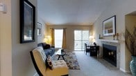 This single-story residence offers a thoughtfully designed layout centered around a welcoming living room that draws the eye upward with its vaulted ceilings. The living area is anchored by a tiled fireplace, adding warmth and character to the space, while soft, plush carpeting underfoot lends a comfortable, inviting atmosphere. Large windows dressed with warm curtains allow natural light to fill the room, and a sliding glass door opens to a private balcony, bringing in views of the surrounding greenery. The open flow between the living area and the adjacent kitchen creates a sense of spaciousness, with the kitchen featuring dark appliances and a functional layout. Wall art and decorative touches throughout contribute to a curated, homey aesthetic. A hallway extends from the main living space, connecting to additional rooms bathed in warm, ambient lighting — suggesting well-proportioned private quarters beyond. The textured ceilings and neutral wall tones throughout provide a versatile backdrop that complements a variety of interior styles. Overall, the space balances comfort and functionality, making it an ideal environment for both relaxed everyday living and casual entertaining.