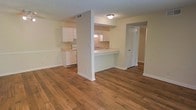 This single-floor unit offers a clean, functional layout designed for comfortable everyday living. The open living area is anchored by rich, warm hardwood-style flooring that extends throughout the space, creating a cohesive and inviting atmosphere. A partial wall with a peninsula-style pass-through counter connects the kitchen to the living area, maintaining an open feel while providing a natural division between the two spaces. The kitchen is outfitted with white cabinetry, white appliances including a range, and a sink positioned at the pass-through, making it both practical and easy to navigate. The light-toned walls and white trim throughout the unit contribute to a bright, airy ambiance. A hallway visible from the main living area leads to additional rooms, suggesting a well-organized floor plan. Ceiling-mounted lighting fixtures provide consistent illumination across the space. The overall design is straightforward and neutral, offering a versatile backdrop that adapts easily to a variety of interior styles and furnishing choices.