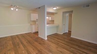 This single-floor unit offers a clean, functional layout designed for comfortable everyday living. The open living area is anchored by rich, warm hardwood-style flooring that extends throughout the space, creating a cohesive and inviting atmosphere. A partial wall with a peninsula-style pass-through counter connects the kitchen to the living area, maintaining an open feel while providing a natural division between the two spaces. The kitchen is outfitted with white cabinetry, white appliances including a range, and a sink positioned at the pass-through, making it both practical and easy to navigate. The light-toned walls and white trim throughout the unit contribute to a bright, airy ambiance. A hallway visible from the main living area leads to additional rooms, suggesting a well-organized floor plan. Ceiling-mounted lighting fixtures provide consistent illumination across the space. The overall design is straightforward and neutral, offering a versatile backdrop that adapts easily to a variety of interior styles and furnishing choices.