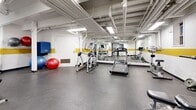 This fitness center offers a comprehensive workout environment spread across a single-level space. The interior is defined by white walls accented with a bold yellow horizontal stripe that runs throughout, lending the space a consistent, energetic aesthetic. The ceiling is exposed, revealing structural beams, ductwork, and conduit overhead, illuminated by fluorescent strip lighting that keeps the area well-lit throughout.

The cardio zone includes treadmills and an elliptical trainer, while a dedicated strength area features a multi-functional cable machine, a barbell station with weight plates, and a substantial rack of hex dumbbells ranging across various weights. Large wall-mounted mirrors expand the visual depth of the space and allow users to monitor their form during workouts.

A functional training corner is stocked with stability balls in multiple sizes, medicine balls on a dedicated rack, a SPRI exercise mat, and various accessories stored on open shelving. Durable rubber flooring covers the entire workout area, providing traction and impact absorption. The layout is open and efficiently organized, accommodating both strength training and cardio exercise without congestion, making it a practical and well-rounded fitness facility.