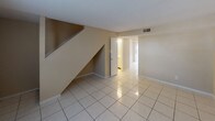 This two-story space is organized across two distinct levels, each serving a clear functional purpose. The main level features light-toned ceramic tile flooring that extends through the open living area and into the kitchen. A staircase rises prominently from the main living space, with the area beneath offering additional utility. The living room receives natural light through a window fitted with horizontal blinds, contributing to a bright and open atmosphere. The kitchen is equipped with warm-toned wood cabinetry, dark countertops, a double-basin sink, and white appliances including a range, dishwasher, and refrigerator, all set on matching tile flooring. A full bathroom on the main level includes a toilet, a single-basin vanity with under-cabinet storage, a lighted mirror, and a separate tub-shower combination with white tile surround. The upper level hosts carpeted bedrooms finished with neutral wall tones and textured ceilings. Each room benefits from natural light via windows with blinds. An additional bathroom upstairs mirrors the clean, functional design of the main-level bath. Throughout, the space maintains a consistent neutral color palette and straightforward layout, offering a practical and livable environment.