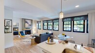 This single-floor residence offers a thoughtfully designed open-concept layout that seamlessly connects the kitchen, dining, and living areas. The kitchen is appointed with white flat-panel cabinetry, stainless steel appliances including a gas range and refrigerator, and a generous peninsula with a built-in sink and bar seating. Pendant lighting above the peninsula adds a refined touch, while light hardwood flooring runs consistently throughout the entire space, enhancing its bright, cohesive aesthetic.

The living area is anchored by a fireplace framed by a clean mantel, and is lined with a bank of black-framed multi-pane windows that allow abundant natural light and offer views of surrounding greenery. A dedicated dining space sits adjacent to additional windows, reinforcing the airy, light-filled character of the home.

The bedroom is a serene and well-proportioned retreat, featuring black-framed windows with garden views, recessed lighting, and ample closet space accessible via a hallway. The bathroom is finished in a clean, minimalist style with a floating white vanity, a round mirror, and a tub-shower combination. White walls throughout the residence create a neutral, versatile backdrop that complements the modern design sensibility of the space.