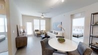 This thoughtfully designed single-floor residence offers a clean, contemporary aesthetic throughout. The open-concept layout seamlessly connects the dining and living areas, creating a welcoming and functional living environment. Light hardwood-style flooring runs throughout the main living space, complementing the neutral, warm white walls that lend the home an airy, expansive feel.

The living area is anchored by a ceiling fan that enhances comfort, while large windows fitted with white horizontal blinds allow generous amounts of natural light to fill the room. A statement abstract artwork adorns one wall, adding a curated touch to the space. The dining area features a round white table paired with upholstered chairs, positioned conveniently adjacent to a dark wood credenza that provides additional storage.

A glimpse into the bedroom reveals a continuation of the bright, serene palette, suggesting a restful private retreat. The open doorway between the living and sleeping areas reinforces the home's sense of openness and flow.

Overall, the space balances modern design with practical comfort — characterized by its neutral tones, clean lines, abundant natural light, and well-considered layout that makes efficient use of every area.