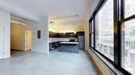 This single-floor space showcases a contemporary open-concept layout that seamlessly integrates the kitchen and living areas. The kitchen is a standout feature, equipped with dark espresso cabinetry that contrasts cleanly against light countertops. A central island provides additional workspace and serves as a natural dividing point within the open floor plan. Stainless steel appliances, including a refrigerator, microwave, and range, lend a modern, functional aesthetic to the kitchen.

The flooring throughout is a polished concrete surface, adding an industrial character that complements the overall design. Track lighting runs along the ceiling, offering flexible illumination across the space. A notable architectural highlight is the large, floor-to-ceiling windows with bold dark frames along one wall, flooding the interior with abundant natural light while offering expansive urban views of the surrounding cityscape. The ceiling height contributes to a generous sense of openness and volume. The neutral color palette throughout creates a clean, versatile backdrop suitable for a variety of uses and furnishing styles.